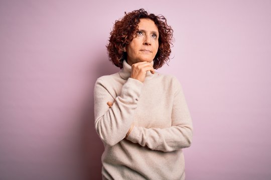 Middle age beautiful curly hair woman wearing casual turtleneck sweater over pink background with hand on chin thinking about question, pensive expression. Smiling with thoughtful face. Doubt concept.