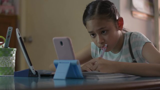 Adorable asian girl watching lesson online from mobile phone and digital tablet during doing homework. Female teenager studying from home with video call during COVID-19 pandemic situation.