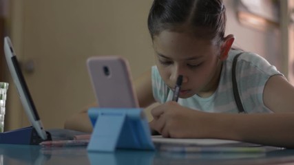 Adorable asian girl watching lesson online from mobile phone and digital tablet during doing homework. Female teenager studying from home with video call during COVID-19 pandemic situation. - Powered by Adobe