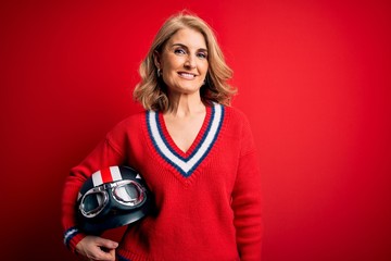 Fototapeta premium Middle age beautiful blonde motorcyclist woman holding moto helmet over red background with a happy face standing and smiling with a confident smile showing teeth