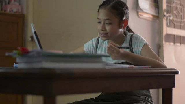 Adorable asian girl wearing earphones and using digital tablet for lesson online at home. Female teenager studying from home with video call during COVID-19 pandemic situation.