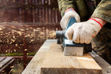 Hands of a joiner with an electric planer. Flying shavings, wooden board. Woodwork. The work of a professional.