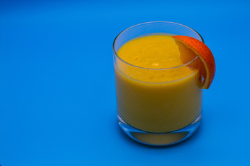 Isolated glass of yellow mango fruit shake with slice of orange with blue background and copy space