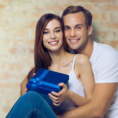 Portrait of beautiful amorous smiling couple holding blue gift box. Love, relationship, dating, happy lovers, concept, over loft style wall. Square composition.