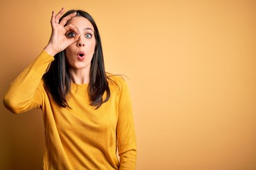 Young brunette woman with blue eyes wearing casual sweater over yellow background doing ok gesture shocked with surprised face, eye looking through fingers. Unbelieving expression.