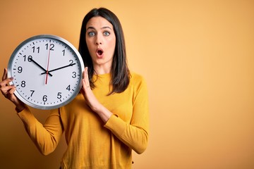Young woman with blue eyes doing countdown holding big clock over yellow background scared in shock...