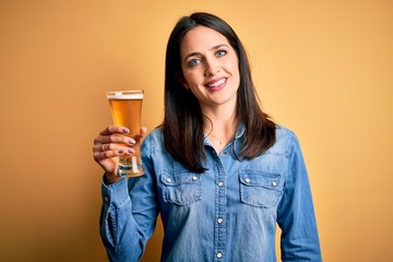 Young woman with blue eyes drinking glass of beer standing over isolated yellow background with a happy face standing and smiling with a confident smile showing teeth