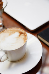 A cup of cappuccino on the table next to a notebook and a laptop, close-up