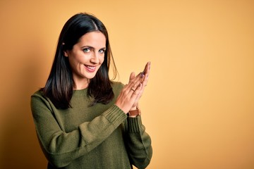 Young brunette woman with blue eyes wearing green casual sweater over yellow background clapping and applauding happy and joyful, smiling proud hands together