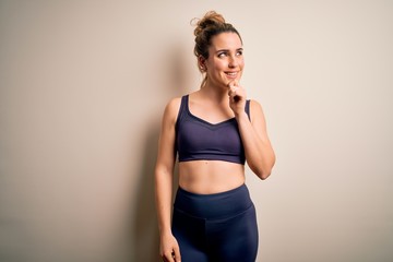 Young beautiful blonde sportswoman doing sport wearing sportswear over white background with hand on chin thinking about question, pensive expression. Smiling with thoughtful face. Doubt concept.