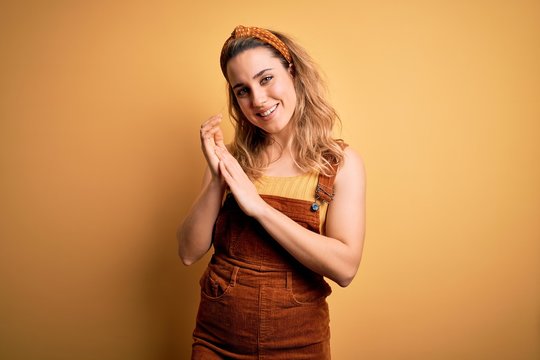 Young beautiful blonde woman wearing overalls and diadem standing over yellow background clapping and applauding happy and joyful, smiling proud hands together