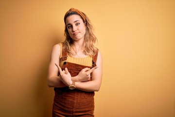 Young beautiful blonde woman wearing overalls and diadem standing over yellow background Pointing to both sides with fingers, different direction disagree