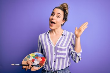 Young beautiful blonde artist woman painting using brush and palette over purple background very happy and excited, winner expression celebrating victory screaming with big smile and raised hands