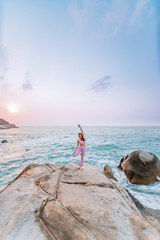 Pregnant woman practices yoga, standing in vrksasana position on the beach, on the rocks. Hands of girl in a pose meditating. Serenity practicing at sunrise, meditation. Woman in pink-purple tracksuit