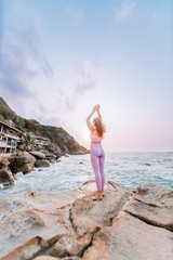 Pregnant woman practices yoga, standing in vrksasana position on the beach, on the rocks. Hands of girl in a pose meditating. Serenity practicing at sunrise, meditation. Woman in pink-purple tracksuit