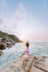 Pregnant woman practices yoga, standing in vrksasana position on the beach, on the rocks. Hands of girl in a pose meditating. Serenity practicing at sunrise, meditation. Woman in pink-purple tracksuit