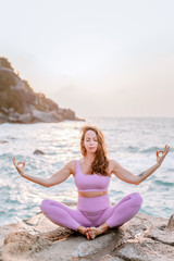 Fototapeta na wymiar Pregnant woman practices yoga, sitting in lotus position on the beach, on the rocks. Hands of girl in a pose meditating. Serenity practicing at sunrise, meditation. Woman in a pink-purple tracksuit.