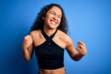 Fototapeta na wymiar Young beautiful woman with curly hair wearing casual t-shirt standing over blue background excited for success with arms raised and eyes closed celebrating victory smiling. Winner concept.
