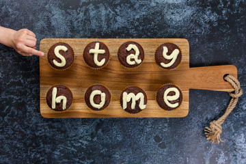 Concept flat lay Stay Home. One childrens hand reach out chocolate muffins, with cream letters, stays on wooden cutting board. Eight cupcakes make up the two words. Family on self-isolation.