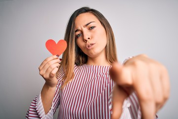 Young beautiful blonde romantic woman holding red paper heart over white background pointing with finger to the camera and to you, hand sign, positive and confident gesture from the front