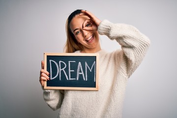 Young beautiful blonde woman with blue eyes holding blackboard with dream word message with happy face smiling doing ok sign with hand on eye looking through fingers