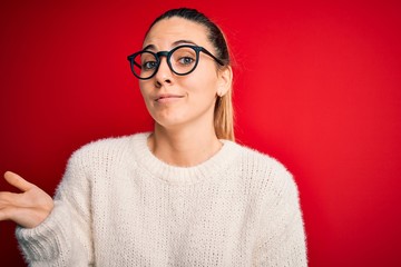 Beautiful blonde woman with blue eyes wearing sweater and glasses over red background clueless and confused expression with arms and hands raised. Doubt concept.