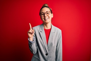 Young beautiful blonde businesswoman with blue eyes wearing glasses and jacket showing and pointing up with finger number one while smiling confident and happy.