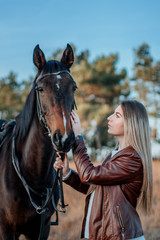 Fototapeta premium Beautiful elegance woman kisses your hokse. Has smiling face, brown leather jacket and hat. Has slim body. Portrait nature. People and animals. Equestrian. Close up.