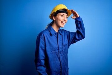 Young beautiful worker woman with blue eyes wearing security helmet and uniform very happy and smiling looking far away with hand over head. Searching concept.