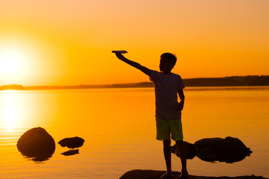 Little boy holds a paper airplane in hand at sunset. A child raised his hand up to sky and plays with origami in the evening at lake. Silhouette