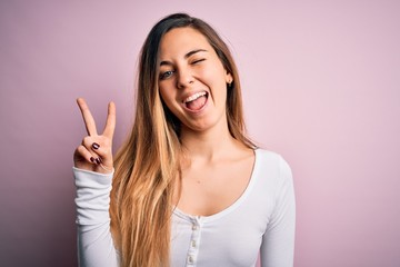 Young beautiful blonde woman with blue eyes wearing white t-shirt over pink background smiling with happy face winking at the camera doing victory sign with fingers. Number two.