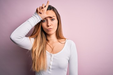 Young beautiful blonde woman with blue eyes wearing white t-shirt over pink background making fun of people with fingers on forehead doing loser gesture mocking and insulting.