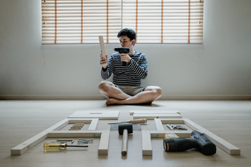 Young asian man self assembly wooden furniture with electric screwdriver as her move into their new...