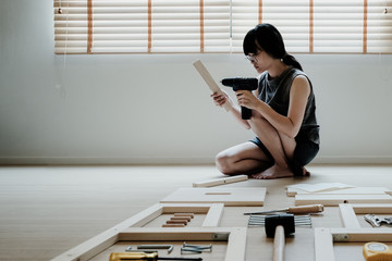 Young asian woman self assembly wooden furniture with electric screwdriver as her move into their...