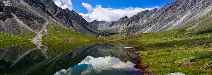 Panorama of a mountain landscape with reflection in the lake, peaks of a mountain range in clouds view from a green valley © Baikal360