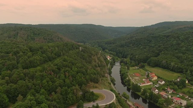 Aerial view of forest, road and river near small houses and village during sunset. Cloudy weather.