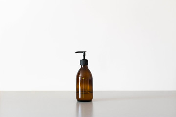 An amber glass refillable soap dispenser or bottle against a plain white background, sat on a cool grey surface.  