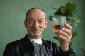 Front view of senior man holding glass of whisky or brandy and looking at camera in loft style room with light green walls and houseplants on background. Alcohol addiction