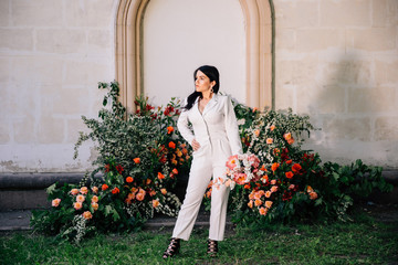 original bride in a white trouser suit posing against the background of an unusual composition of fresh flowers in coral color for an outdoor wedding ceremony