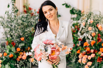 original bride in a white trouser suit posing against the background of an unusual composition of fresh flowers in coral color for an outdoor wedding ceremony