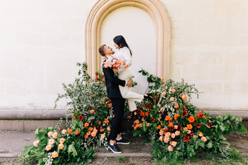 the groom is circling the bride in his arms on the background of an unusual composition of natural flowers of the color coral for a wedding ceremony