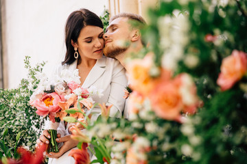 the bride and groom gently posing between floral arrangements of fresh flowers in color coral created for an outdoor wedding ceremony