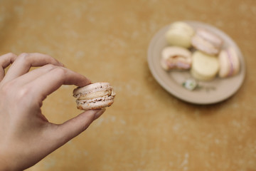 Yellow homemade macarons with blossom are lying on the brown plate on the golden background. Person is holding a beige macaron in a hand.