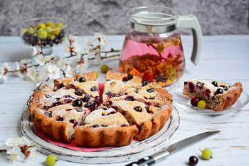  Appetizing pie with blackcurrant, blueberries and icing sugar, herbal tea in a transparent teapot, a branch of blossoming apricot and a slice of cake on a plate on a light wooden table.