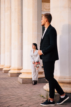 Bride And Groom In A White Pantsuit Posing On The Background Of Large White Columns