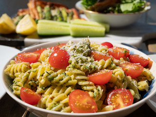 Fusilli Pasta with pesto, chopped cherry tomatoes and grated parmesan cheese. Placed on a wooden table outdoors. Background of salad, cucumber batons and sliced lemon. 