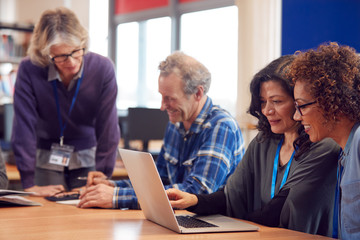 Teacher With Group Of Mature Adult Students In Class Sit Around Table And Work In College Library