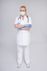 full length portrait of female doctor in surgical mask, glasses and gloves posing over gray