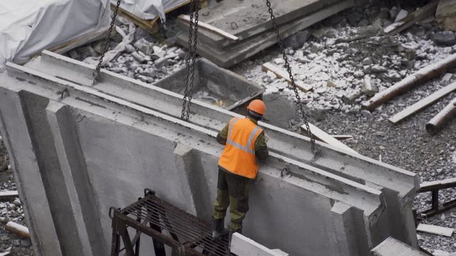 The builder works at the construction site of the house - he hooks the concrete slab with chains, the crane lifts the slab