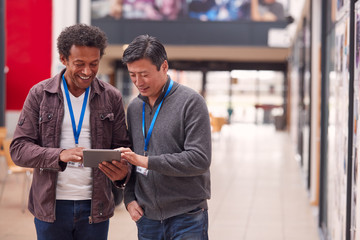 Two Mature Male Students Or Teachers With Digital Tablet Walk Through Hall Of College Building